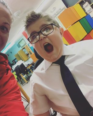 friendships | Muscular Dystrophy News | Max is wearing a tie and glasses, mouth wide open, in front of what appears to be a classroom. Half-pictured is his mom, Betty, in red.