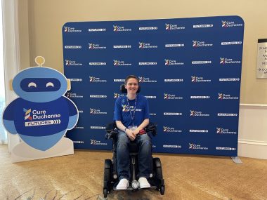 CureDuchenne | Muscular Dystrophy News | Hawken poses in front of a CureDuchenne Futures backdrop and a blue robot with the organization logo.
