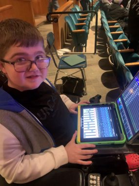 A 17-year-old boy with Duchenne muscular dystrophy sits in his power chair at the back of an auditorium. He's holding an iPad on his lap to work light and sound for the show choir's performance.