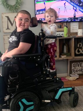 A young girl stands perched on the back of her brother's power wheelchair in the family's living room. The boy, wearing glasses and a black T-shirt, glances back toward her. The girl looks at the camera with her mouth agape. A basketball game is on the TV in the background.