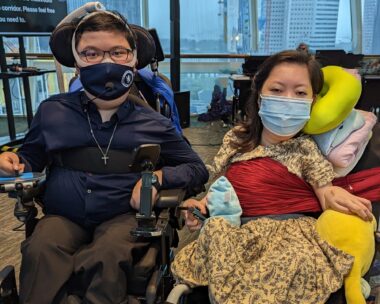 Two people face the camera, seated in wheelchairs in what appears to be a high-rise building with a wall of windows. The man on the left wears brown pants, a dark blue shirt, a cross around his neck, glasses, and a dark blue mask. The woman on the right wears a golden dress with brown designs, with a red sash across her chest. She also wears a light blue mask.