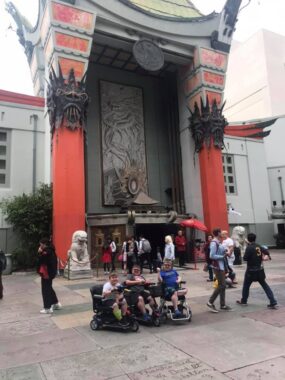 Three boys, all seated in mobility scooters, smile for a photo in front of the TCL Chinese Theatre in Los Angeles.