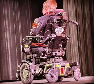 The back of a power wheelchair with a white magnet with lettering near the top at center. A male is in the chair; we see the back of his short reddish-blond hair, the suggestion of glasses, and what appears to be a leather jacket with a red patch on the left shoulder. He is facing long stage curtains.