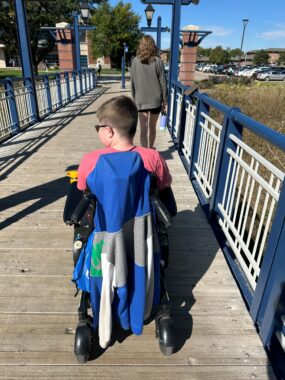 A young man in a wheelchair, his back to the camera, moves along a wooden bridge with blue and white metal guardrails. He has close-cropped brown hair and is wearing sunglasses and a light red shirt; a blue and gray jacket is on the back of his chair. A woman walks some yards ahead of him, and the end of the bridge is in sight, with a parking lot in the distance on the right.