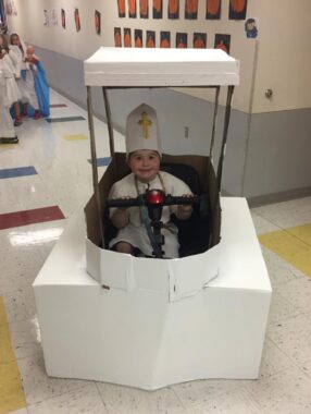 An elementary-aged boy sits on his mobility scooter in the middle of a school hallway. The scooter, however, is barely visible, as it's been covered by a custom-made cardboard structure that looks like a small palanquin. The boy appears to be dressed as a pope, with a tall white mitre hat that features a gold cross. He's sporting a big smile. 