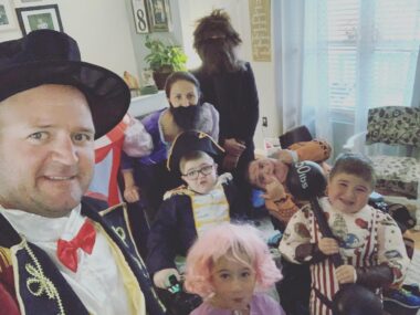 A family selfie, taken by the father, features him, his wife, and five of their kids. All are dressed as various circus characters, and several of the kids are making funny faces. They've grouped together for the photo and appear to be in their living room.