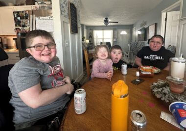 Four children sit at a long , brown kitchen table. Clockwise from left: a teenage boy wearing glasses and a gray T-shirt with red and white lettering; a smiling toddler girl with dark blond hair and a pink top; a young boy with his mouth wide open for the camera and wearing a black T-shirt; and last, another teenage boy wearing glasses and a black T-shirt. Cans of soda and other items are on the table; the kitchen and house in the background have white walls.
