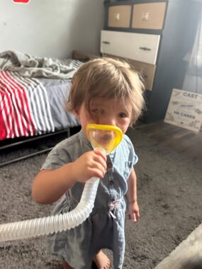 A toddler girl with blond hair and a light gray outfit holds a face mask with yellow edges to her face; the mask is attached to a plastic hose. She's in a bedroom where the rug is gray, a bedspread behind her is gray with sections of red and white stripes, and a dark gray chest has brown and white drawers.