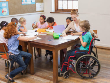 Children, one in a wheelchair, sitting around a table with a teacher.