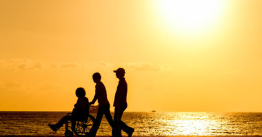 Silhouettes of wo people walking with a person in a wheelchair next to water at sunset