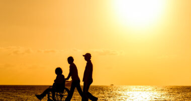 Silhouettes of wo people walking with a person in a wheelchair next to water at sunset