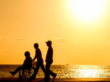 Silhouettes of wo people walking with a person in a wheelchair next to water at sunset