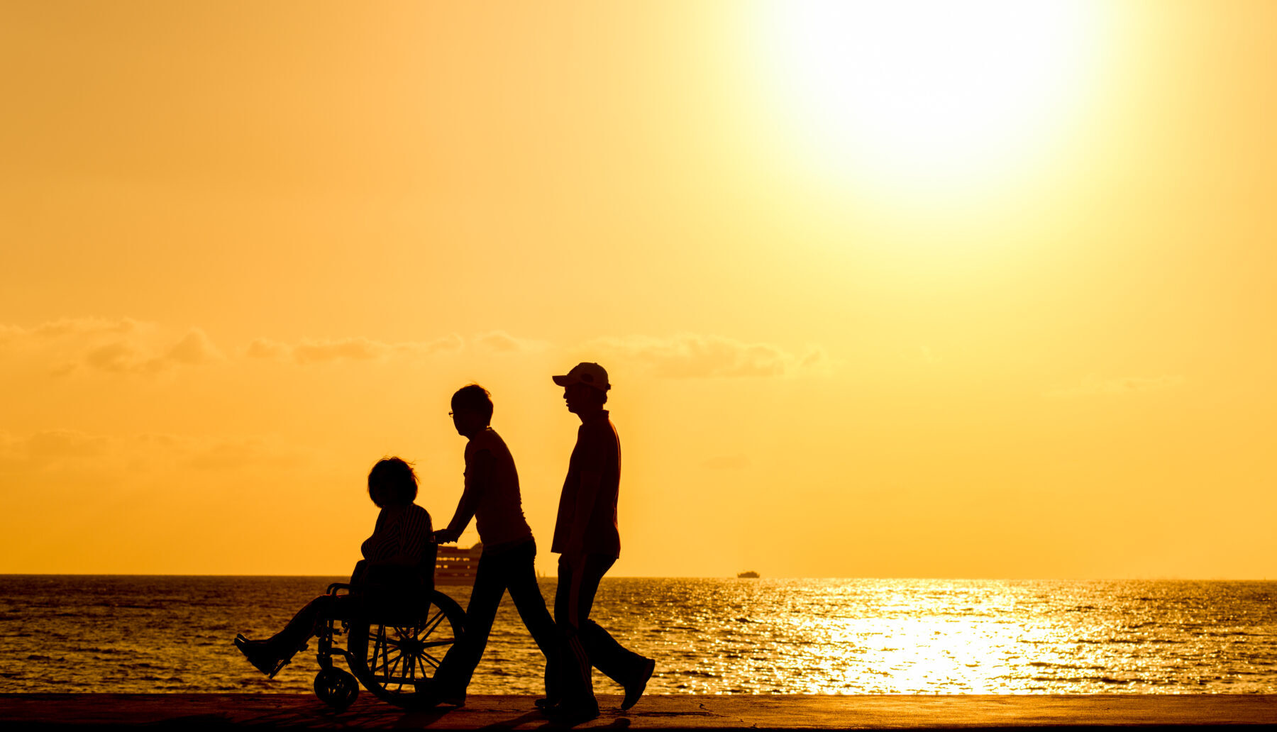 Silhouettes of wo people walking with a person in a wheelchair next to water at sunset