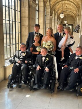 In a hall of windows and arches, eight people, including children, pose for a photo. The three boys in the front rows are sitting in wheelchairs. Behind them are four people, a man holding a little girl.