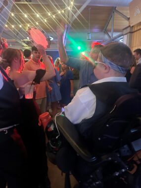 A young man sits in a wheelchair with his back to the camera. A woman with a raised tambourine stands nearby, along with others. A string of lights is tied to the ceiling rafters.