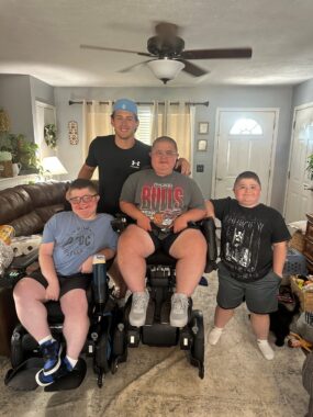 Four males — teenagers and young men — are gathered in a family area. The two in front are in wheelchairs; the one at center is much higher than the other. One young man stands behind them wearing a black shirt and what appears to be a blue baseball cap. The fourth teenager, of short stature, stands at right, wearing a black T-shirt, dark blue shorts and what appear to be white socks.