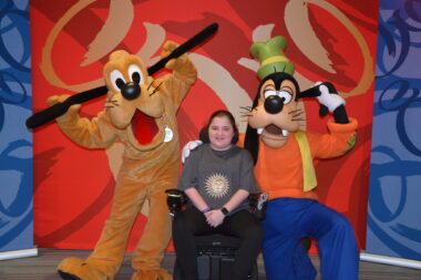 A 16-year-old girl, seated in her power wheelchair, smiles for a photo with Pluto and Goofy at Disney World. They're posing in front of a red and blue backdrop.