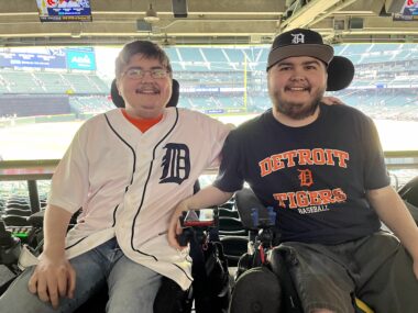 Two men seated in wheelchairs are shown at a baseball stadium.