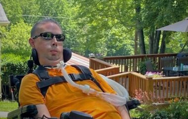 A man outdoors, apparently strapped into a neck support, is in a wheelchair and using what appears to be a breathing tube. He wears sunglasses; behind him is an attractive uncovered porch and lots of greenery.