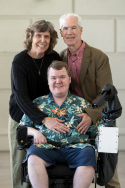 A man in a tropical green shirt is in the center of the photo, flanked by his parents behind him. The mother, to our left, has her right hand on the gentleman in the center, who is in a wheelchair, while the father, to our right, has his left hand on his son's chest.