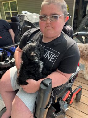 A teenager with close-cropped hair wears a black T-shirt and pale green shorts as he sits in a wheelchair, holding a black-haired puppy with a bit of white on his chest. They're on a wooden deck outdoors, with a partly seen person and dog in the background. 