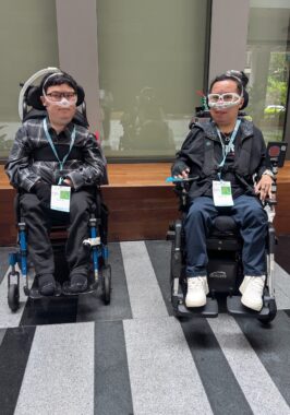 Two men pose in front of a glass wall in a tightly-cropped vertical photo. Both men are in wheelchairs and have ventilators.
