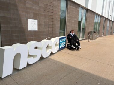 Brayden Cream sits in his power wheelchair outside the Rath Eastlink Community Centre after his college graduation, beside a sign that reads “NSCC Class of 2025.”