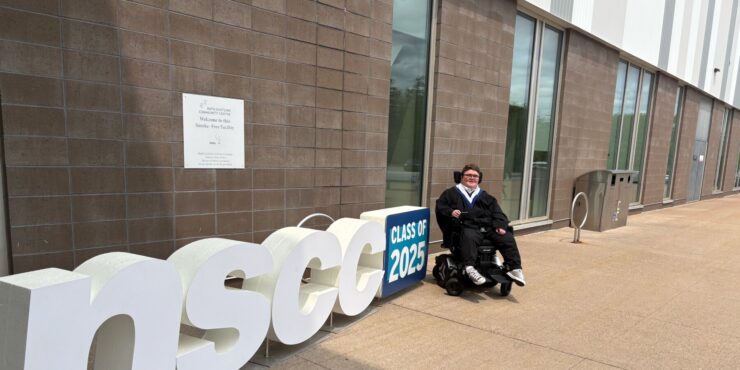Brayden Cream sits in his power wheelchair outside the Rath Eastlink Community Centre after his college graduation, beside a sign that reads “NSCC Class of 2025.”