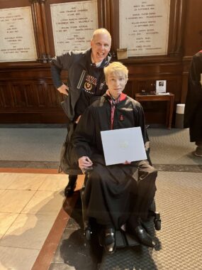 A father and son smile proudly in Harvard gear. The son, in a wheelchair, has a Harvard gown and holds up a degree. 