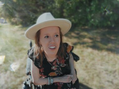 A woman wearing a wide-brimmed hat sits in her power wheelchair in a grassy park.