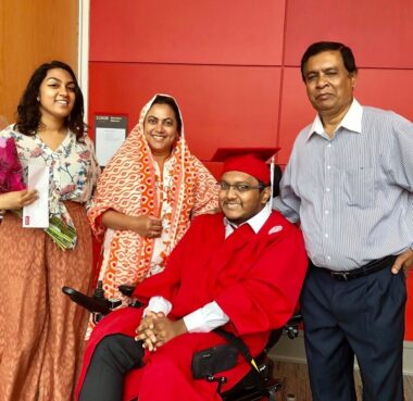 A family of four smiles for a photo at their son's college graduation. He is seated in a power wheelchair and is wearing a red cap and gown, while his sister and parents stand proudly behind him.