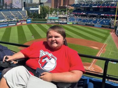 A man in a power wheelchair is seen with a professional baseball stadium behind him.