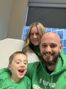 A man and woman lean in for a photo with their 5-year-old son, who's sitting on a large exam chair in a medical office. All three are smiling and wearing green.