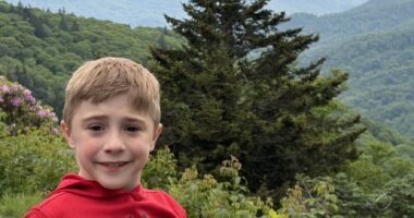 An 8-year-old boy shown from the shoulders up smiles for a photo while standing outside. There are green hills and trees in the background, and it appears to be a foggy day.