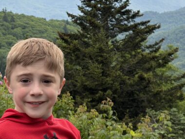 An 8-year-old boy shown from the shoulders up smiles for a photo while standing outside. There are green hills and trees in the background, and it appears to be a foggy day.