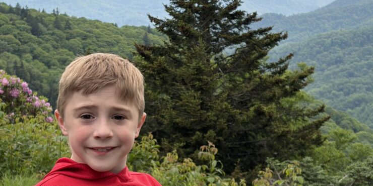 An 8-year-old boy shown from the shoulders up smiles for a photo while standing outside. There are green hills and trees in the background, and it appears to be a foggy day.