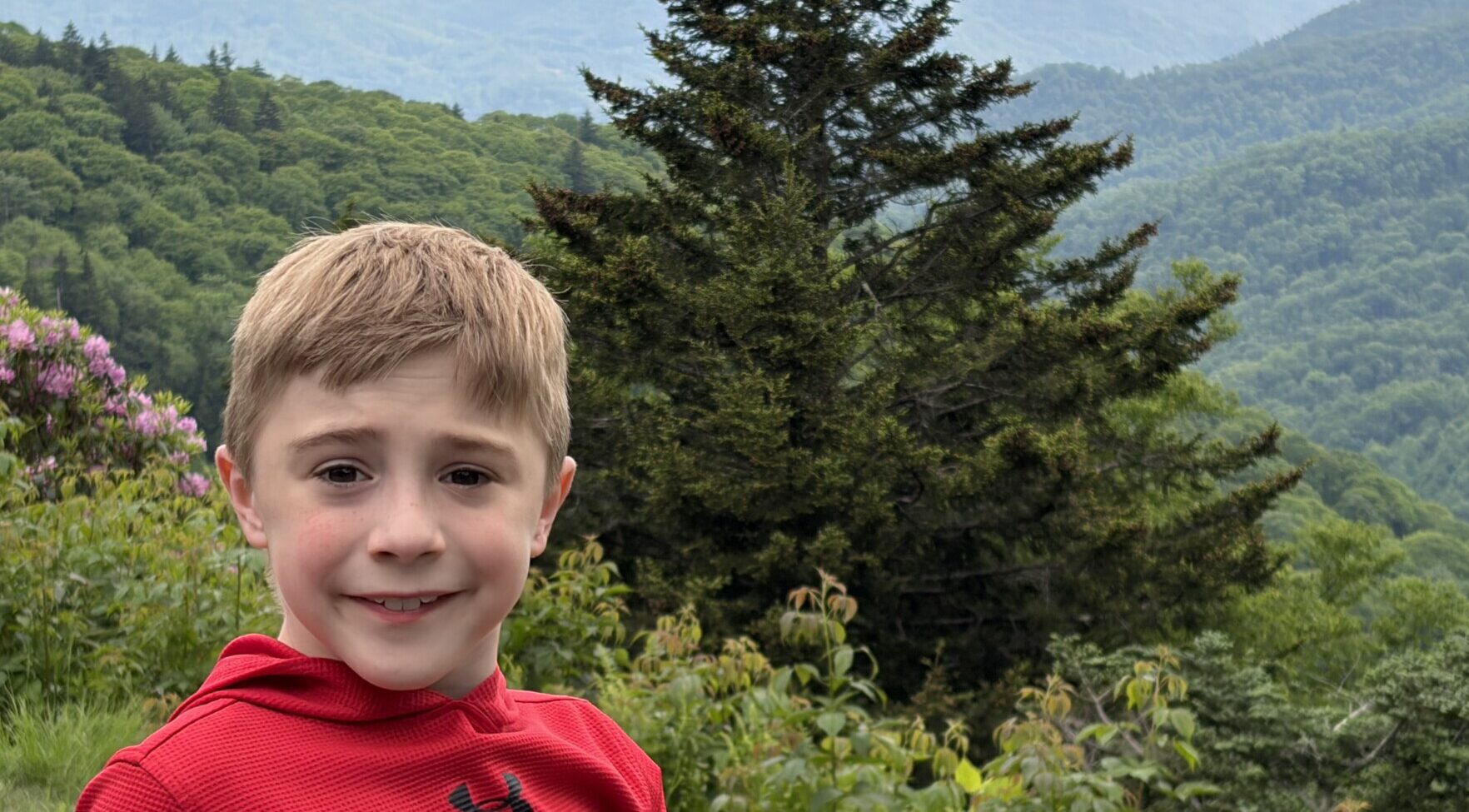 An 8-year-old boy shown from the shoulders up smiles for a photo while standing outside. There are green hills and trees in the background, and it appears to be a foggy day.