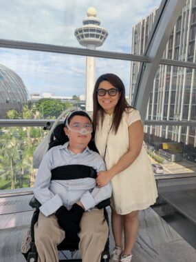 A young man is seated in a wheelchair next to a young woman, who is standing beside him and holds his left arm with her left arm. Behind them is the control tower of an airport. 